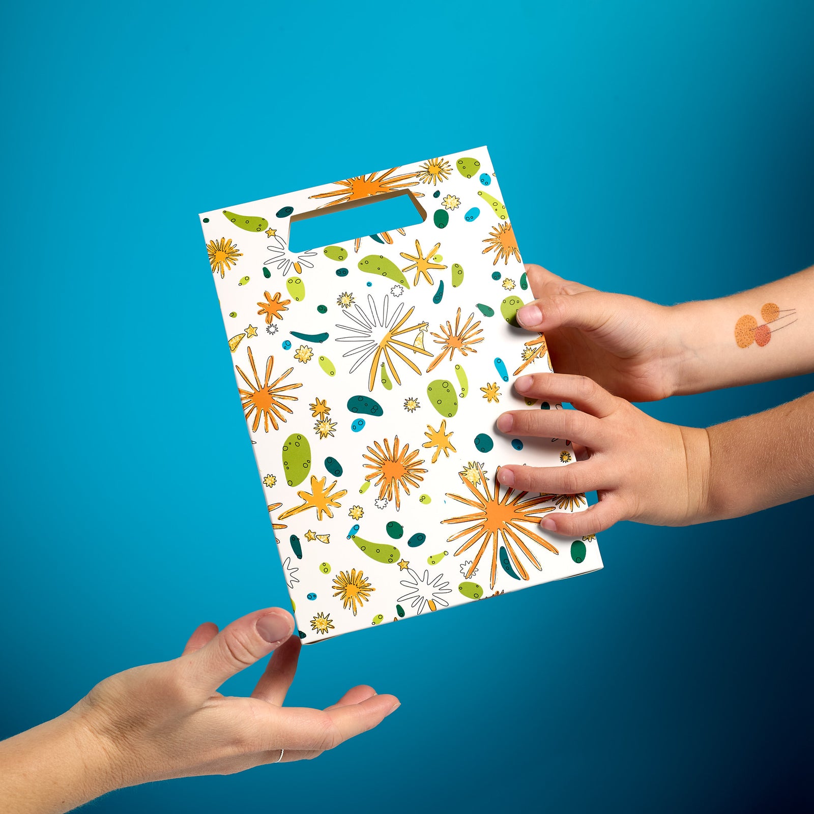 Hands exchanging an ODIDOMI goodie bag with a vibrant, festive design on a blue background. The bag features a playful pattern with gold accents, perfect for kids' party favors and celebrations.