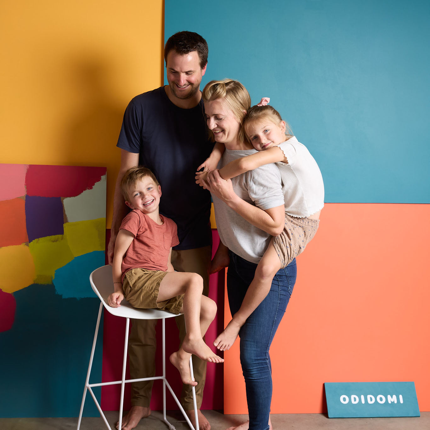 The founders of ODIDOMI, a family-run brand, pose in front of a colorful backdrop. ODIDOMI designs and produces in-house goodie bags and party favors, ensuring high-quality, handcrafted products for kids’ celebrations. A branded wooden sign is displayed on the floor.