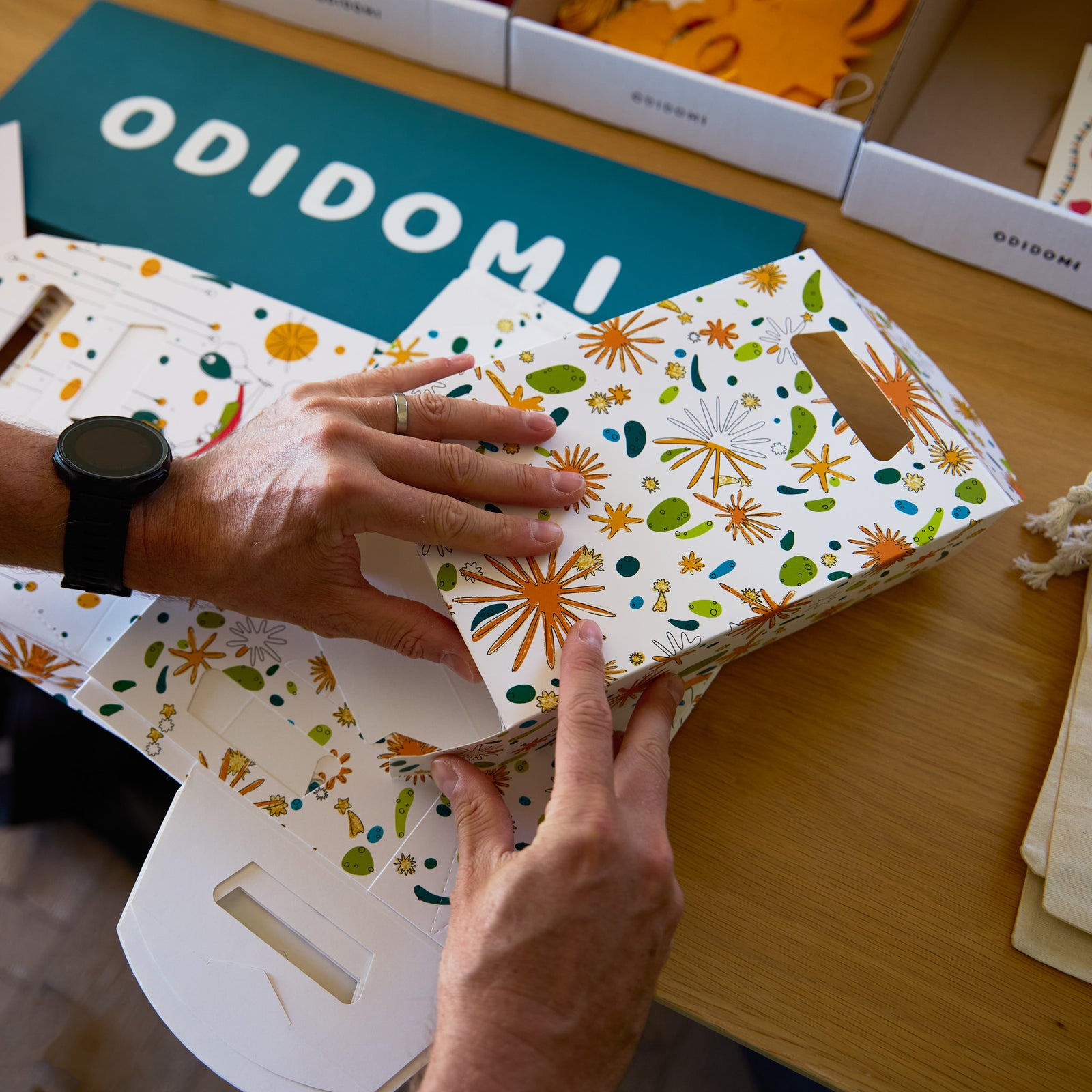 Hands assembling an ODIDOMI goodie bag with a colorful, handcrafted design. ODIDOMI, a family-run brand, designs and produces high-quality, in-house party favor bags for kids' celebrations. A branded wooden sign is visible in the background.