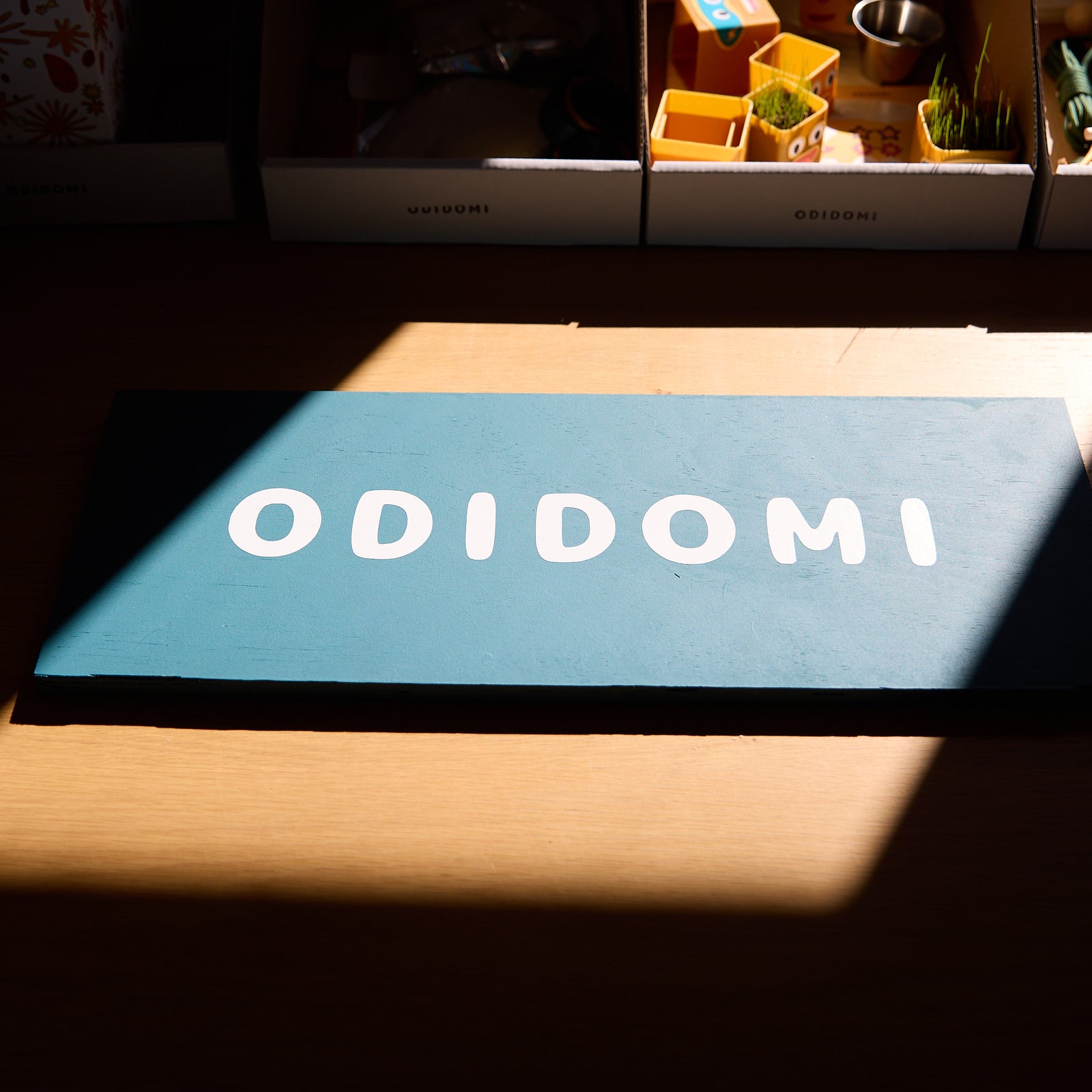 ODIDOMI wooden sign with white lettering on a teal background, placed on a wooden surface with sunlight casting shadows. In the background, ODIDOMI’s curated goodie bags and creative party favor kits are visible, showcasing the brand’s thoughtful packaging and playful designs.